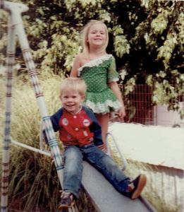 young boy sits on a slide, his young sister behind him in a garden fairy outfit. 